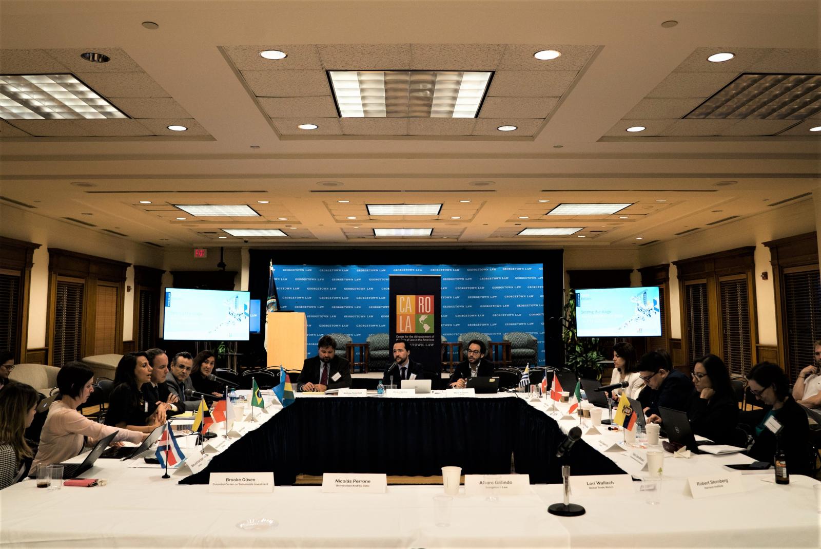 Representatives of Latin American governments, universities, and NGOs talk around a large square table in a room at the Georgetown Law Center. Name tags, country flags, and microphones are set out on the tabletop in front of the representatives.