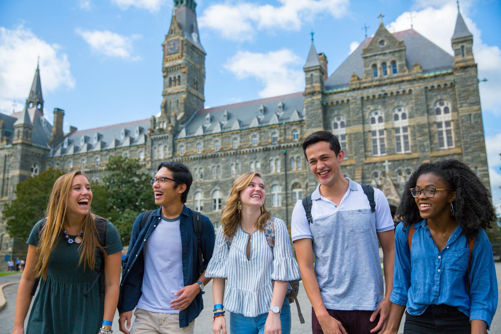 Five undergraduate students walk together and laugh with Healy Hall in the background.