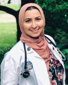 A woman in a white coat and pink hijab smiles