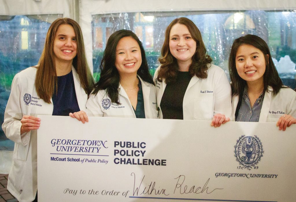 four women in lab coats stand with a check