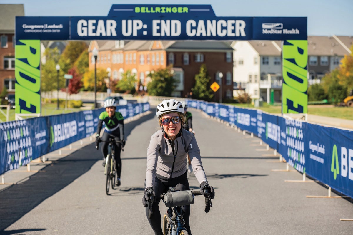 a woman rides her bike to the finish line of the BellRinger ride, past a Gear Up, End Cancer sign