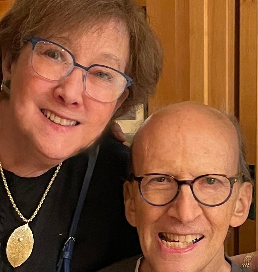 a man with glasses and a woman in a black shirt with yellow necklace smile at the camera