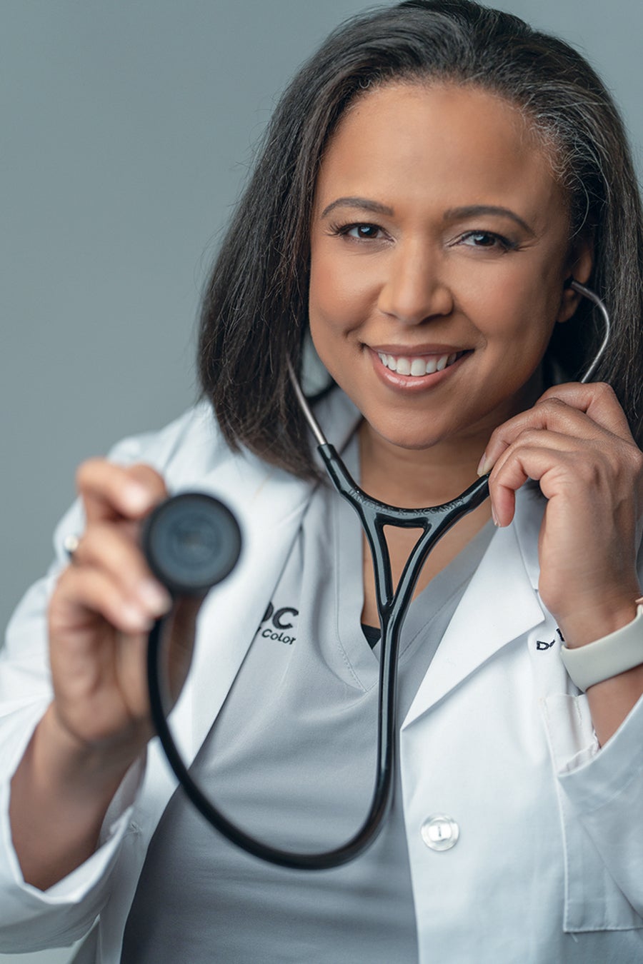 an African American woman wears a white medical coat and holds up a stethoscope