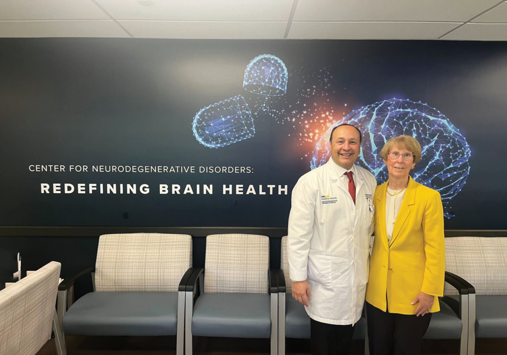 a doctor in a white coat and a woman in a yellow blazer stand in front of a sign for the Center for Neurogenerative Disorders