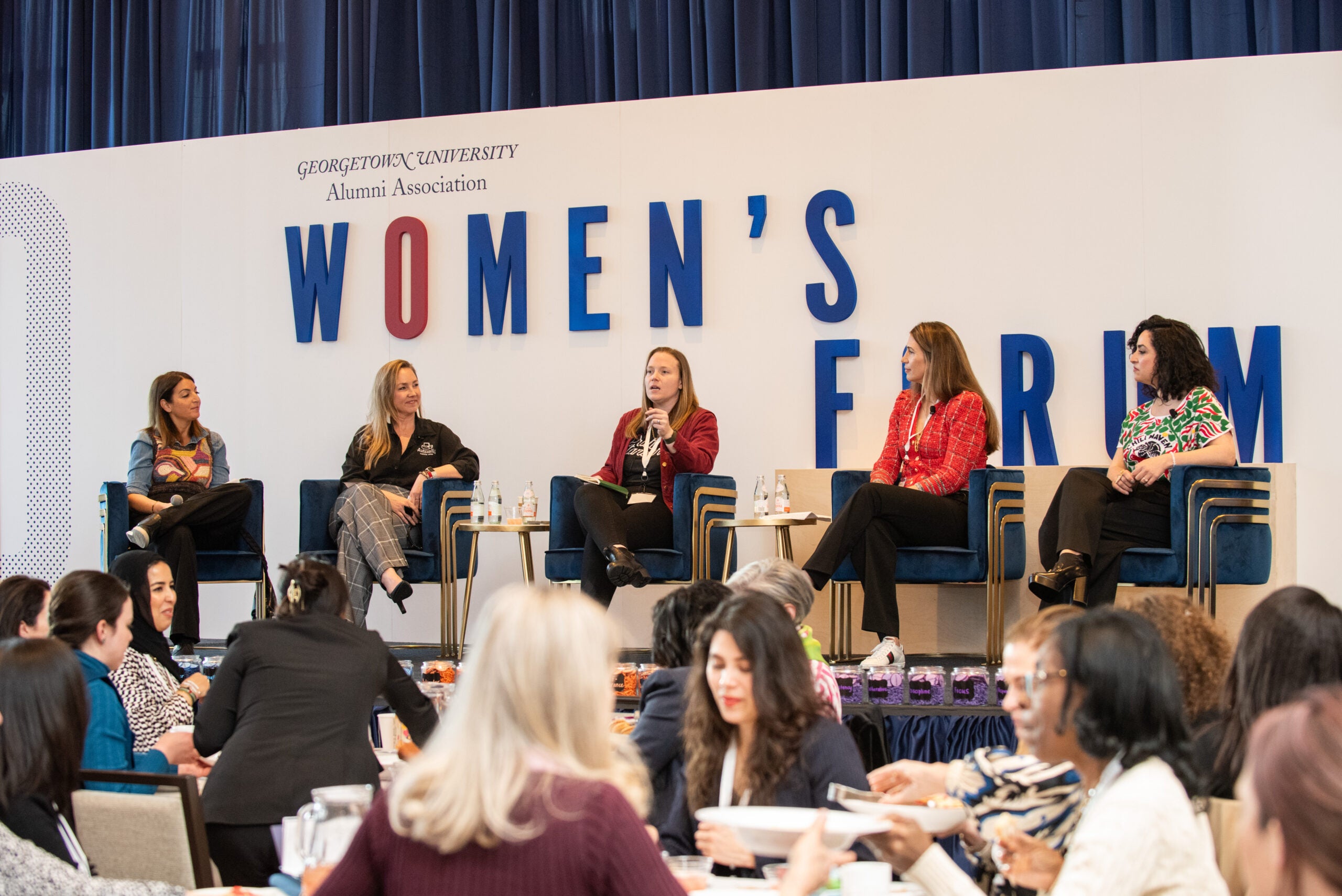 women sit on a stage in front of a crowd, and a background behind them that says "women's forum"
