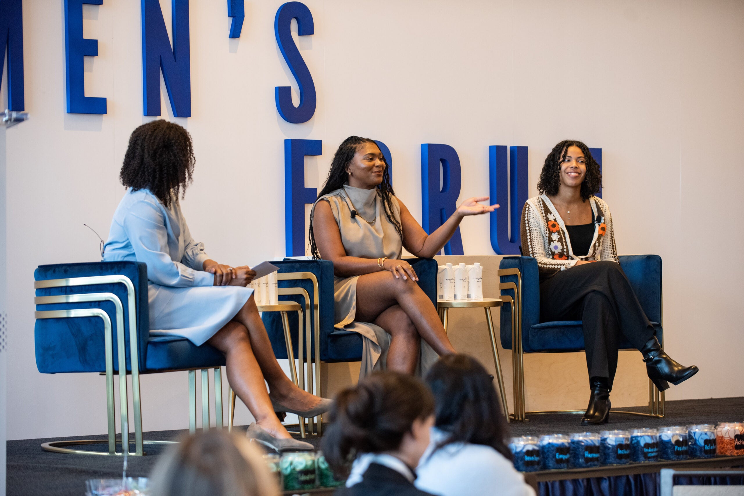 three women sit in blue armchairs on a stage in front of the words "women's forum"