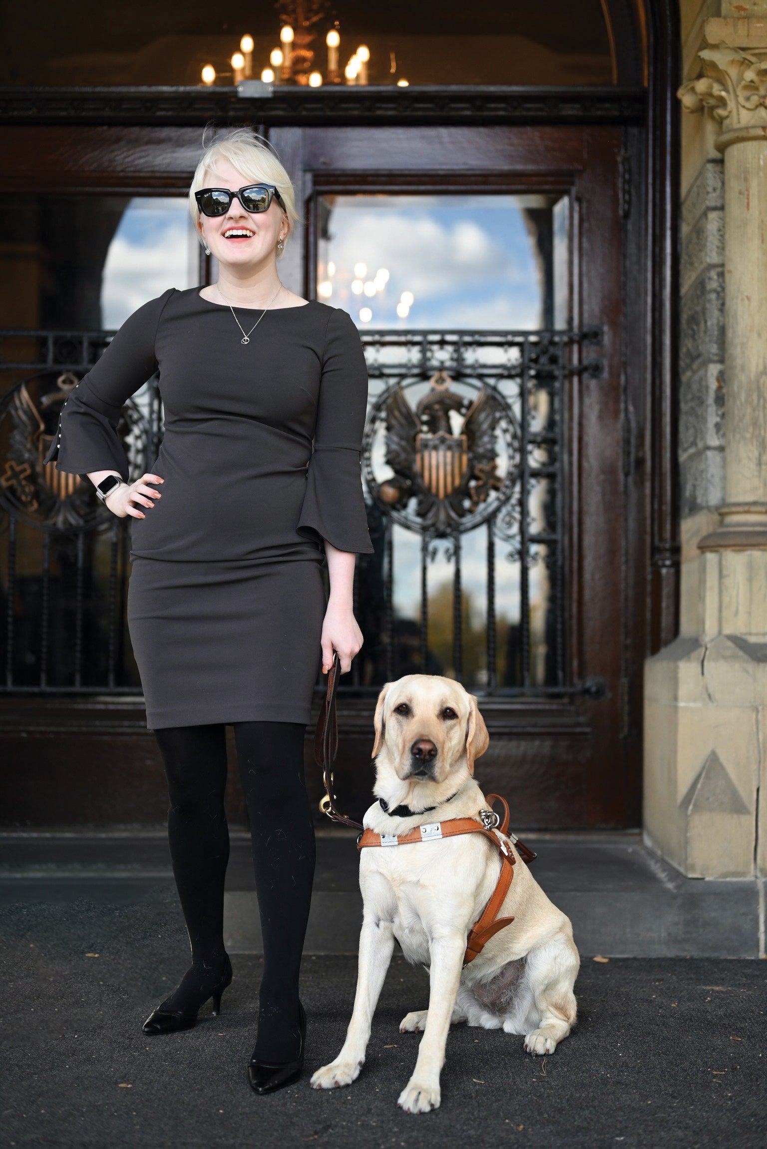 a woman stands outside healy hall doors in a black dress and sunglasses with a golden retriever