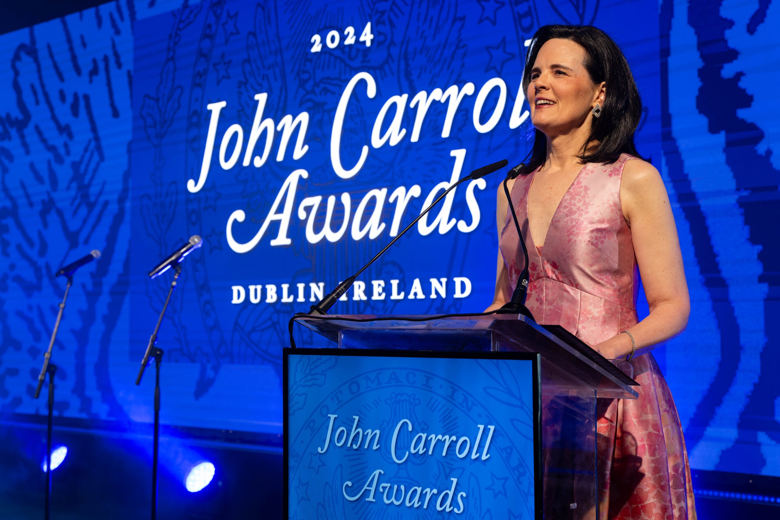 a woman in a pink dress stands at a podium in front of a screen with the words "2024 John Carroll Awards Dublin Ireland"