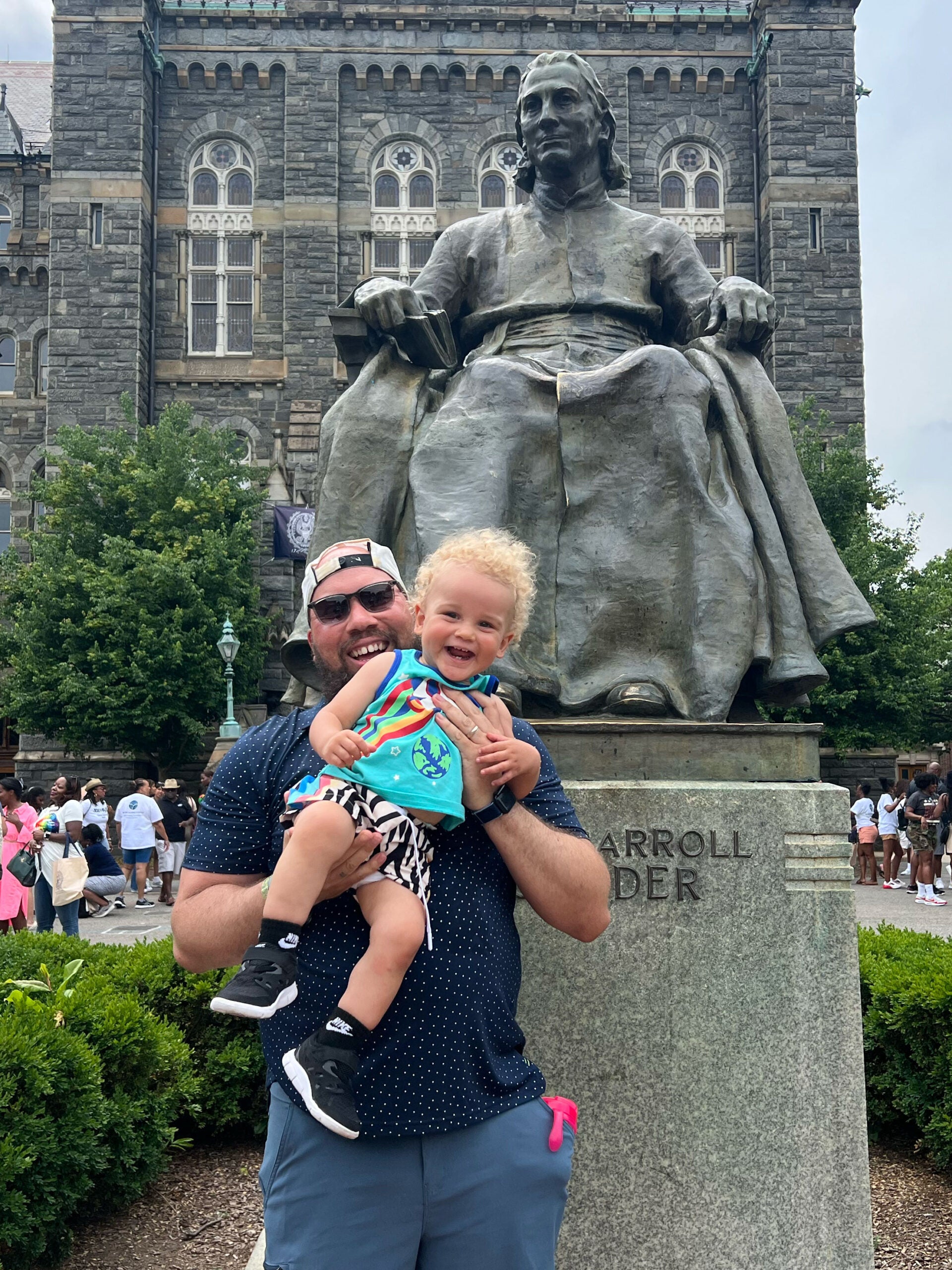 a happy man holds his son in front of the John Carroll statue