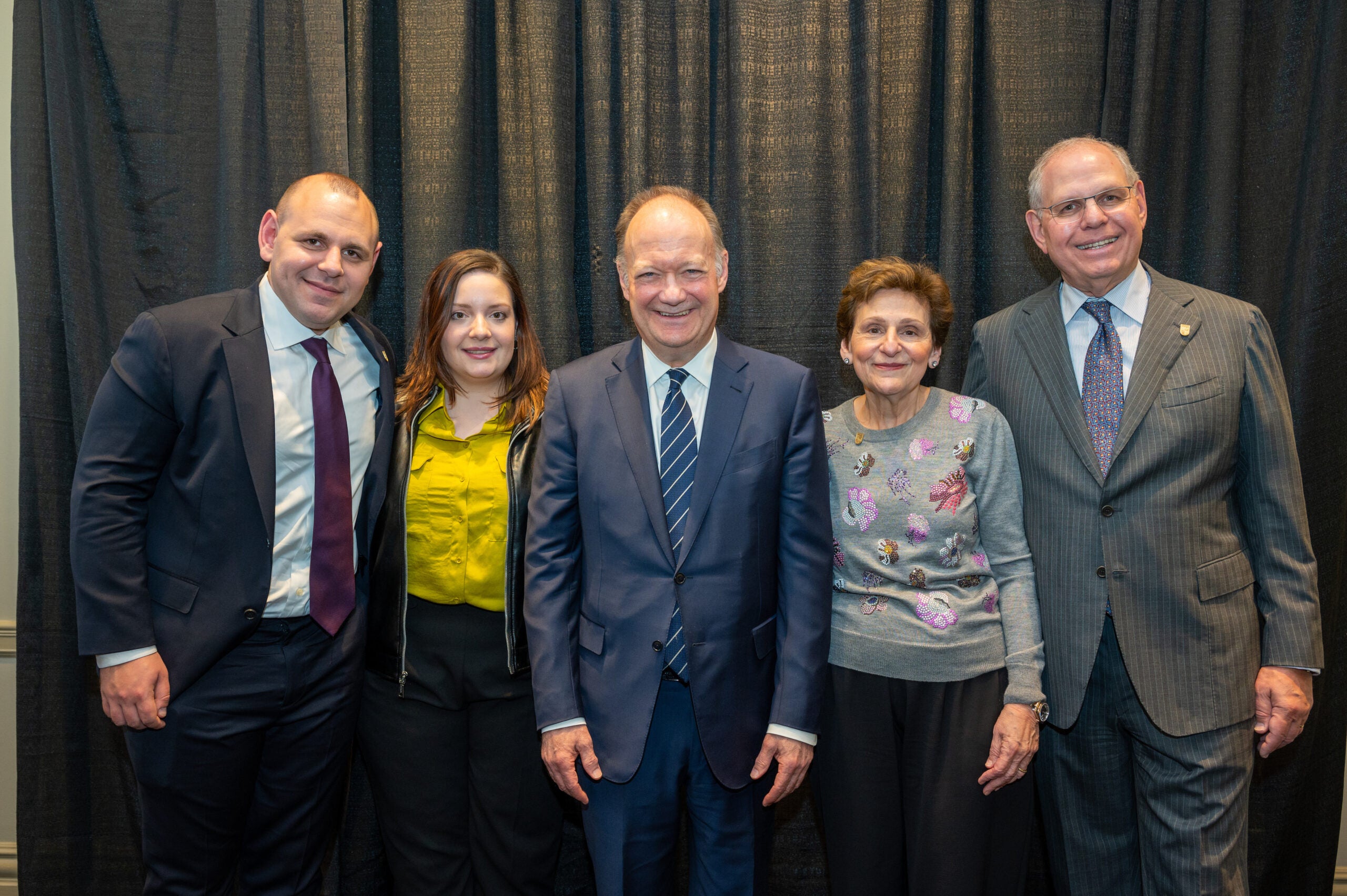 five people (three men, two women) stand together in formalwear. 