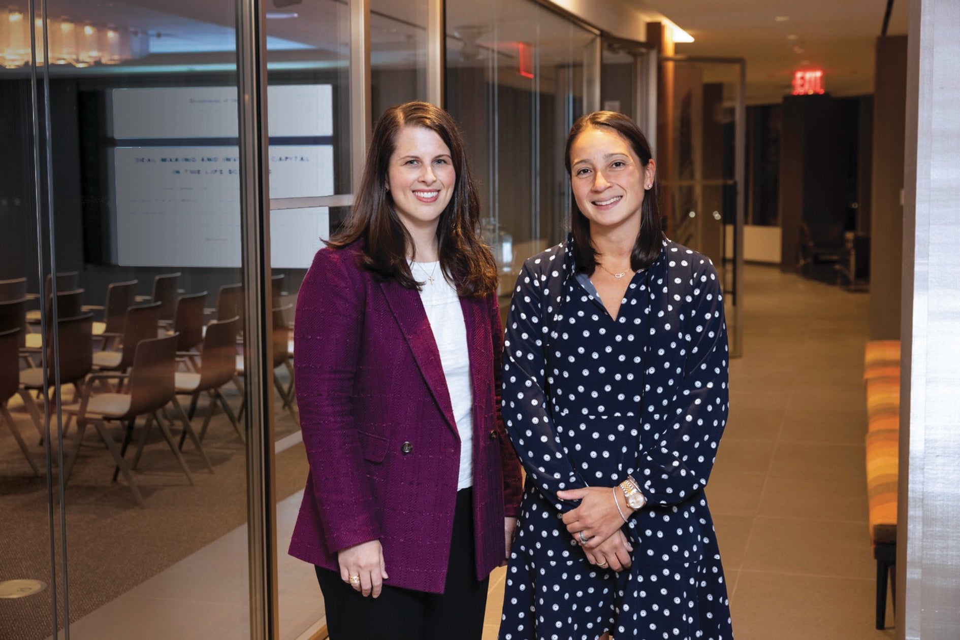 two women stand by a conference room and smile 