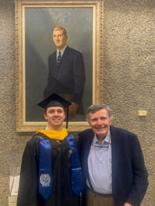 a man in a suit and a young man in a graduation robe stand under a painting of a man