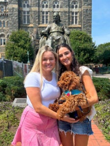 two women stand with puppy in front of a statue on the Georgetown campus