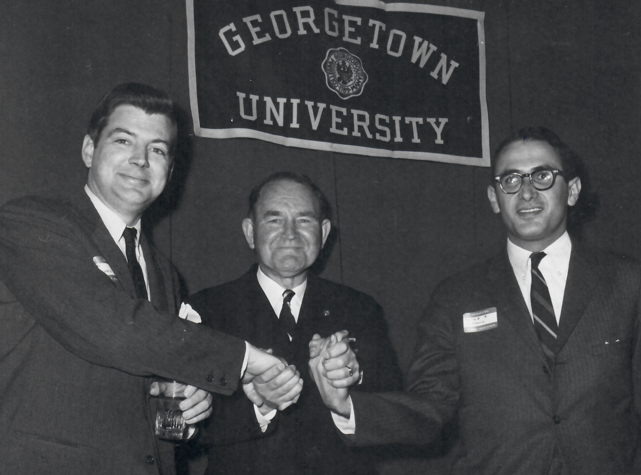 Three men shaking hands in front of Georgetown University banner.