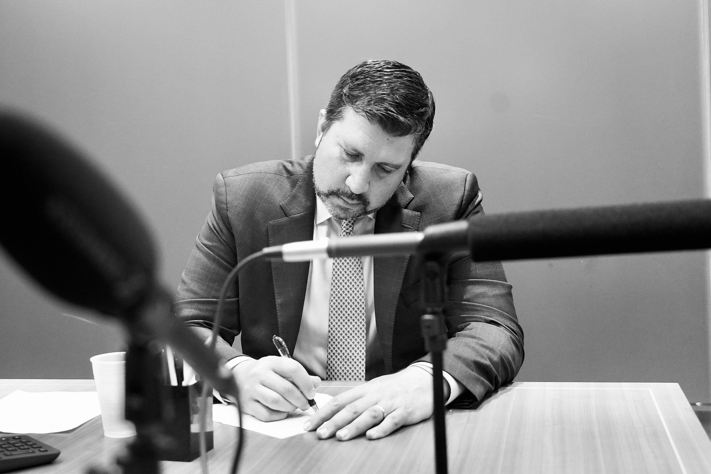 a man in a suit examines a document at a table in front of a microphone