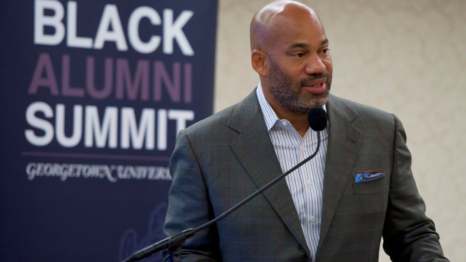 a man in a gray suit stands at a podium in front of a banner that says 