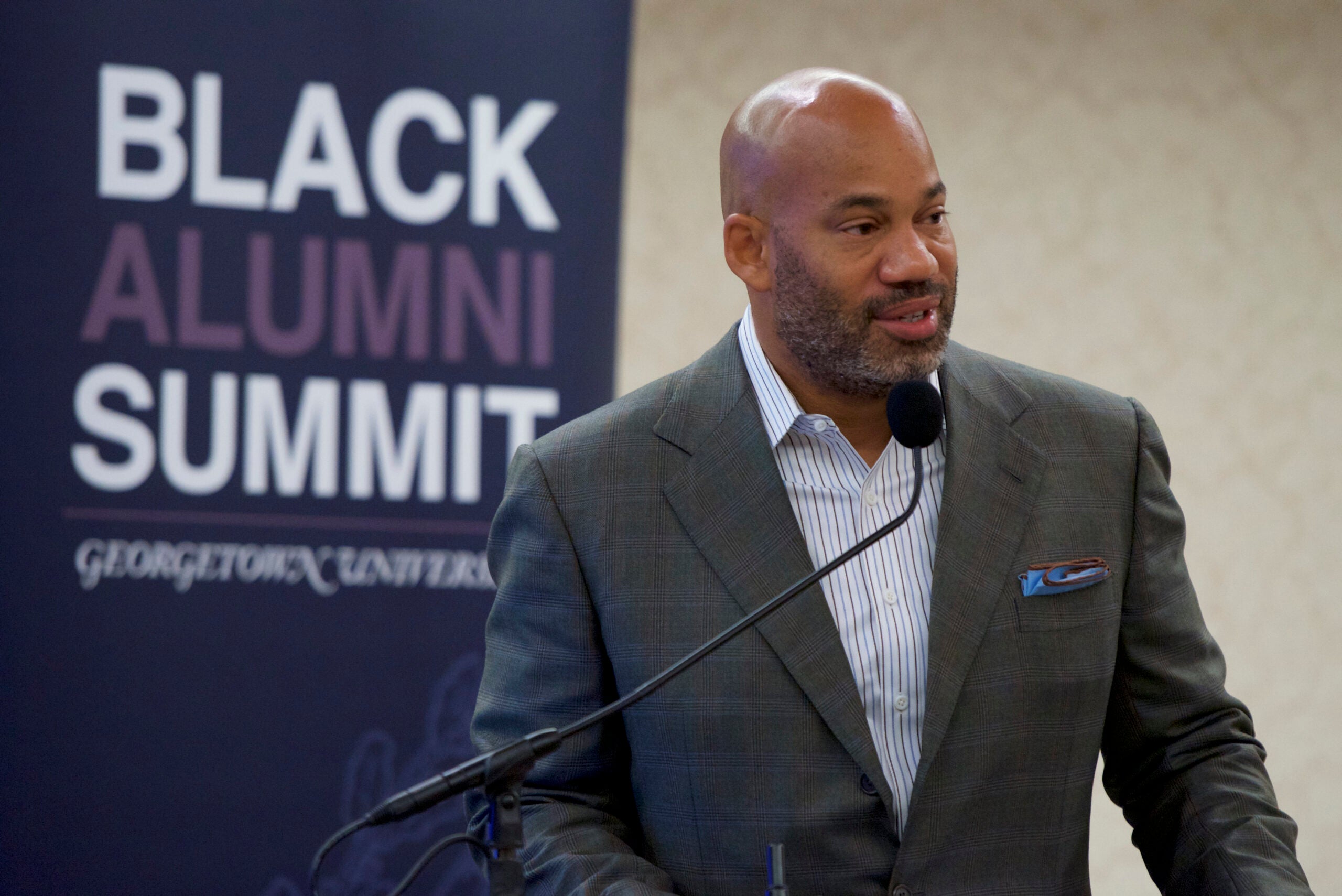 a man in a gray suit stands at a podium in front of a banner that says "Black Alumni Summit: Georgetown University" 