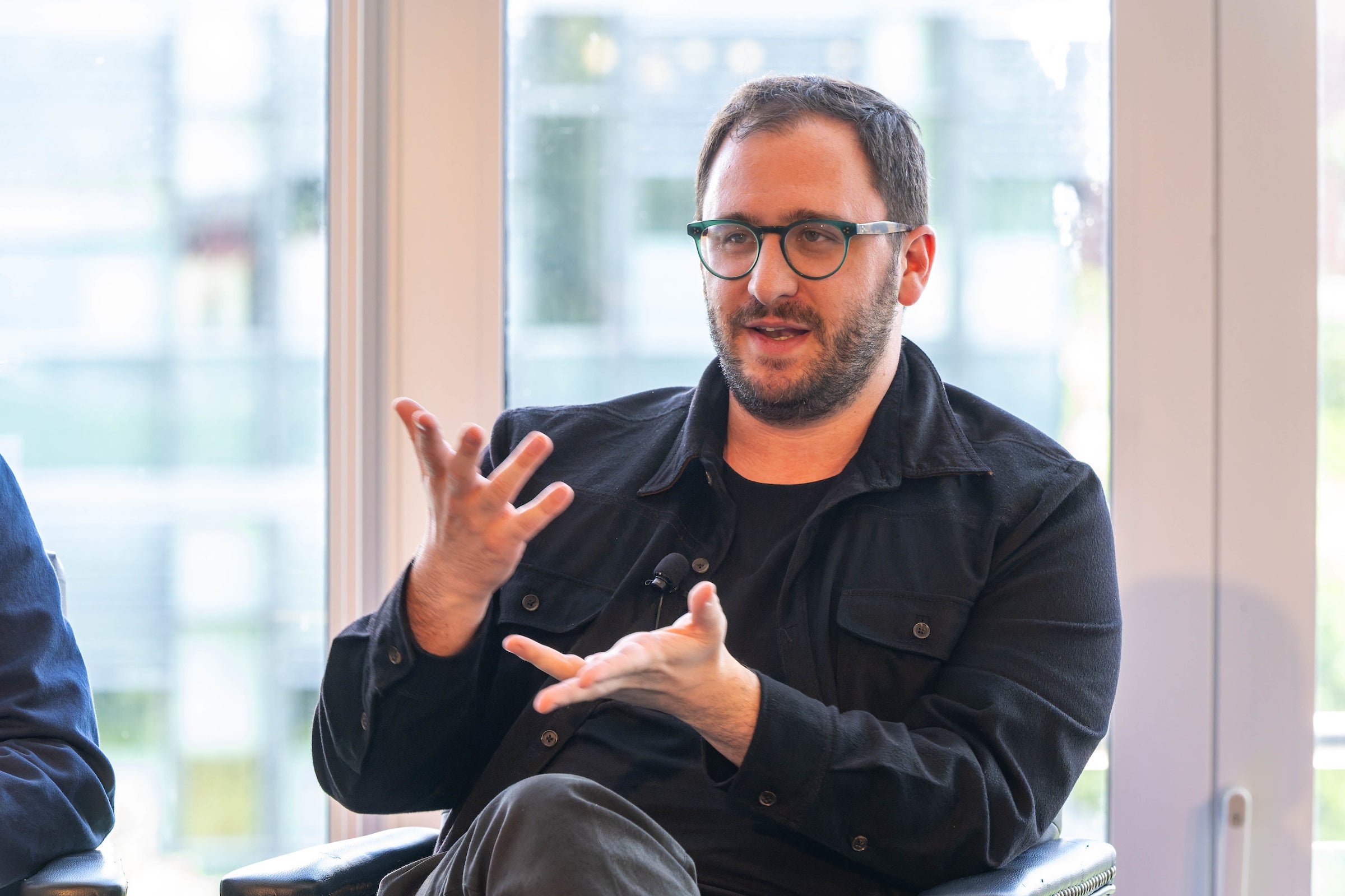 while sitting, a man with glasses gestures with his hands