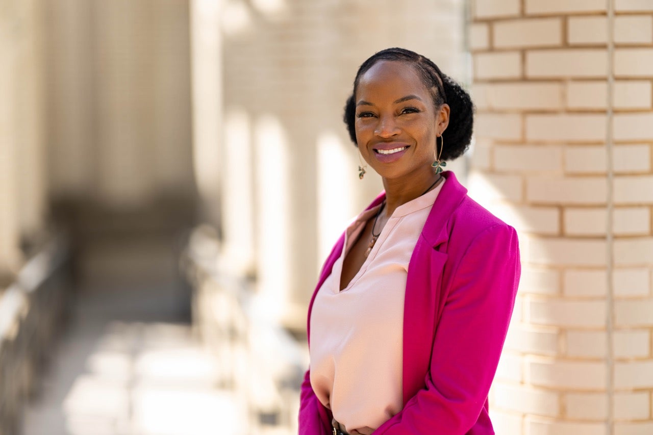 a woman in a magenta blazer and rose shirt outside a brick building