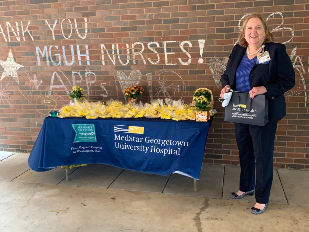 a woman stands at a table of cookies in front of the words "Thank you MGUH Nurses!" on a brick wall