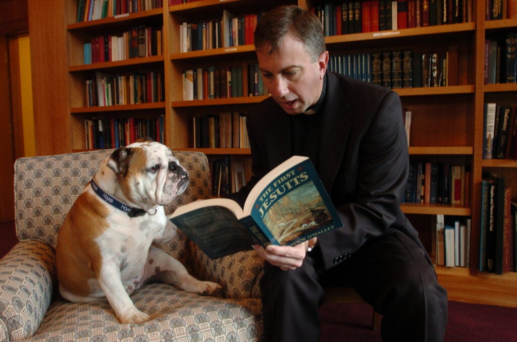 a man read a book to a bulldog seated next to him 
