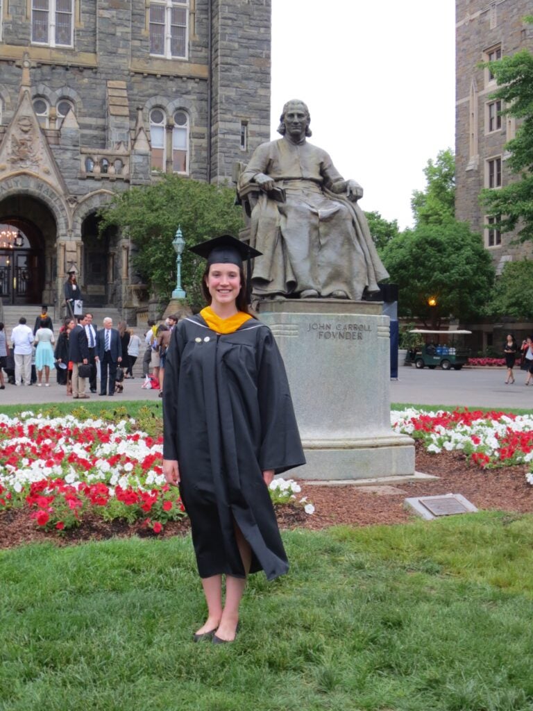 a woman in a graduation gown stands on a campus lawn next to a statue