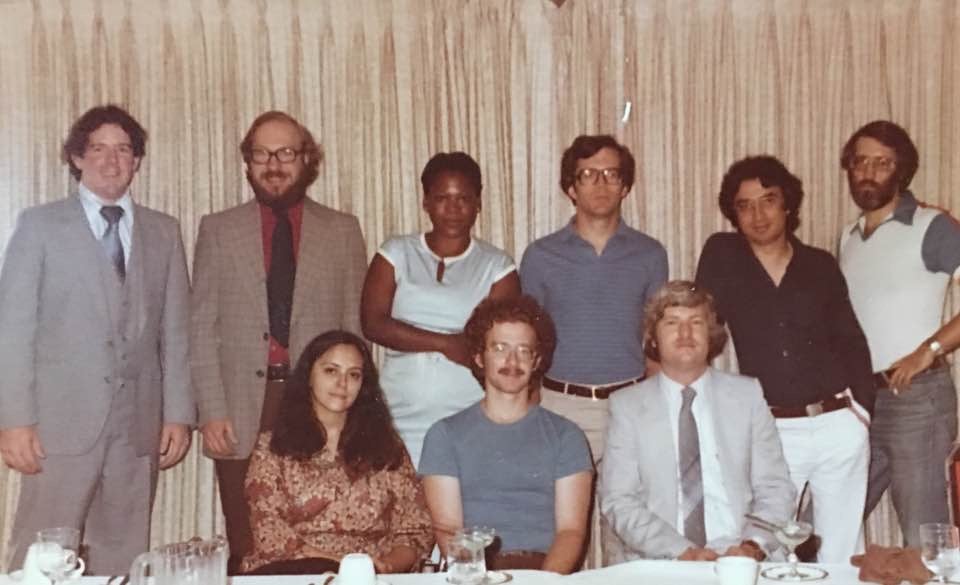 Carmen Ortiz Larsen poses, sitting, for a picture with her collaborators. Two other men sit. Five men stand in the back, and one woman.