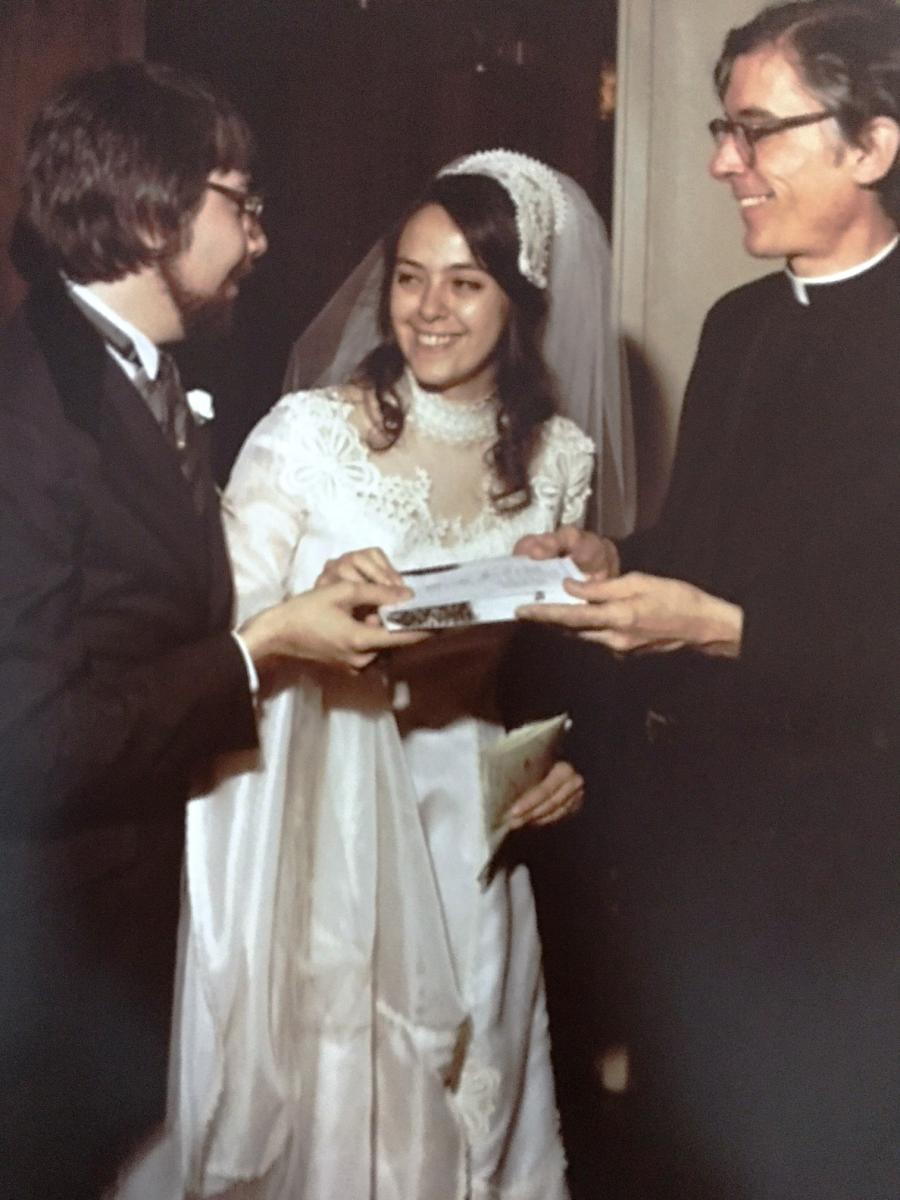 Jan Larsen, at left, in a suit, holds a piece of paper with Carmen Ortiz Larsen, center, in a wedding dress, and Rev. Thomas M. King, S.J., at right, during a wedding ceremony. All three people are smiling.