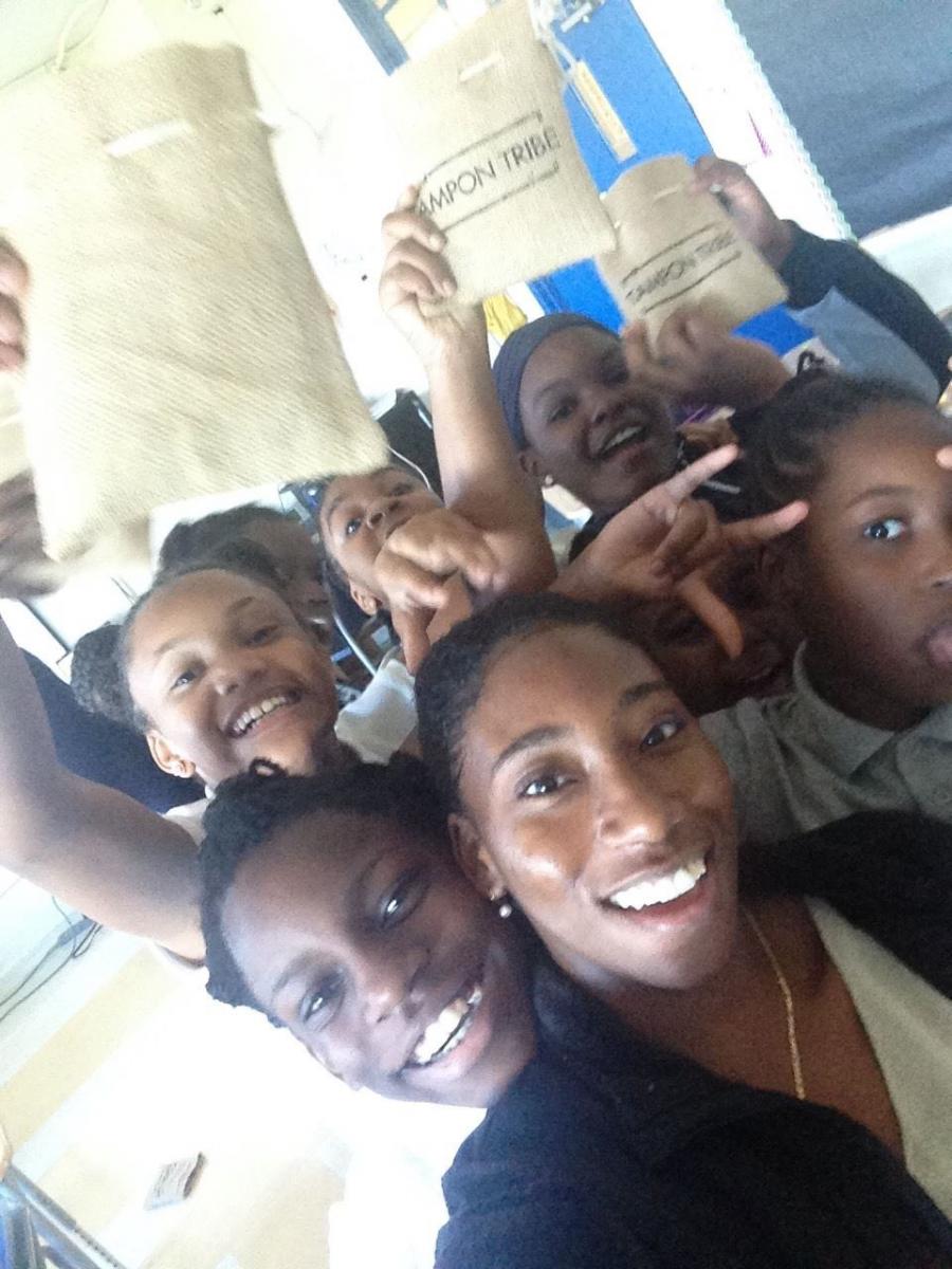 Young women of color in a middle school classroom take a smiling group selfie while holding up burlap pouches emblazoned with the words Tampon Tribe.