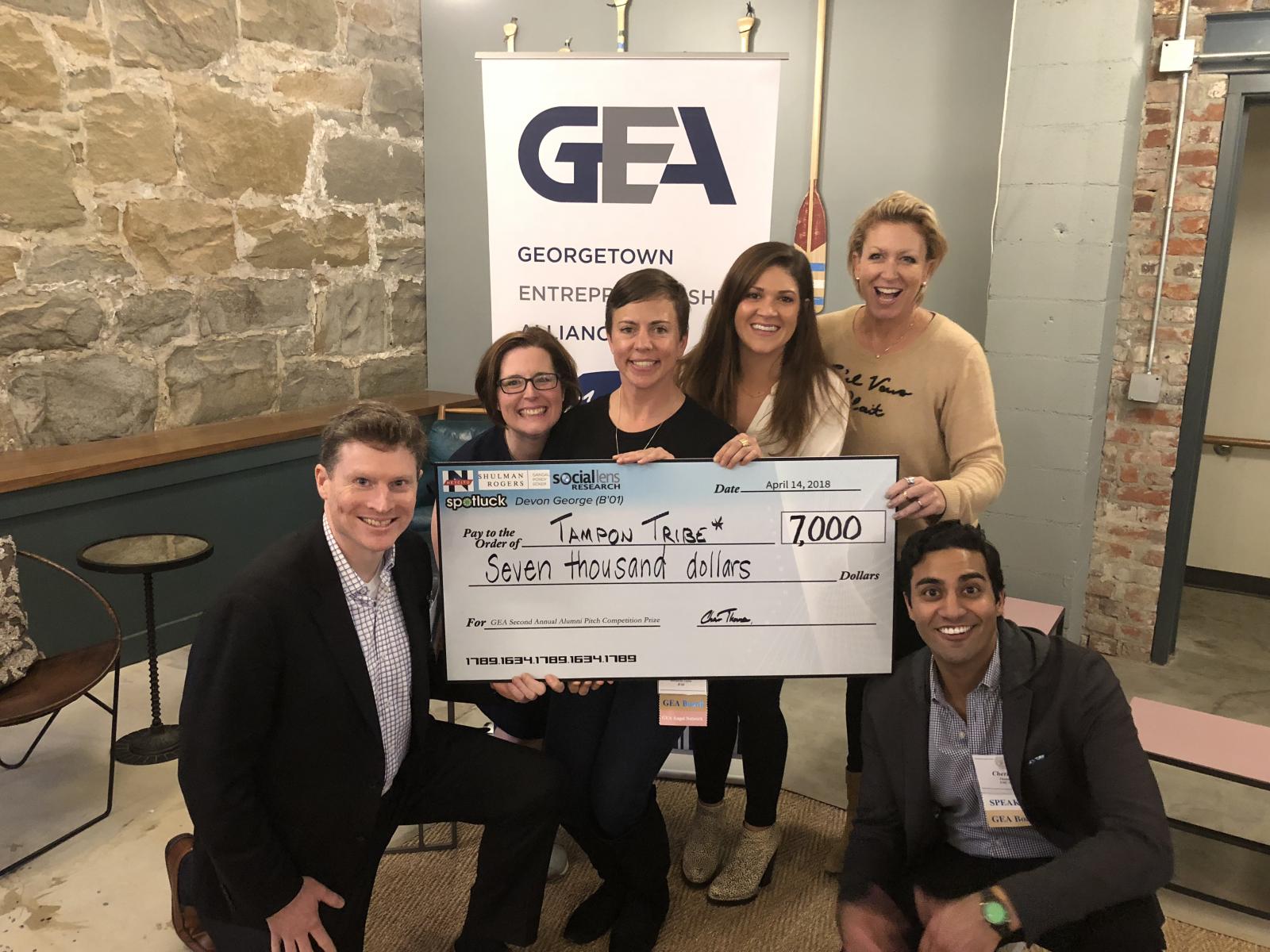 A group of six people in business attire including competition judges Giana Korth and Jennifer Eden pose with a large check for seven thousand dollars made out to Tampon Tribe. Giana Korth holds the check in front of a poster saying Georgetown Entrepreneurship Alliance.
