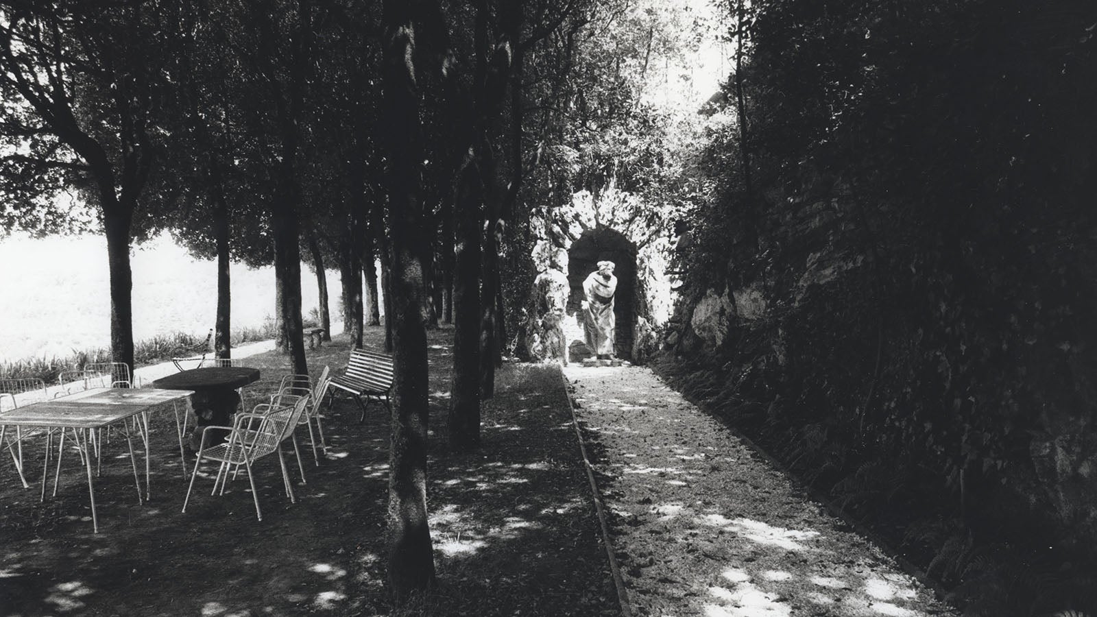 Black and white photo of wire tables and chairs, a stone table, and a bench sitting underneath a grove of trees. To the right, a white pebble pathway leads to a grotto, inside which stands the stone statue of a man wearing robes.