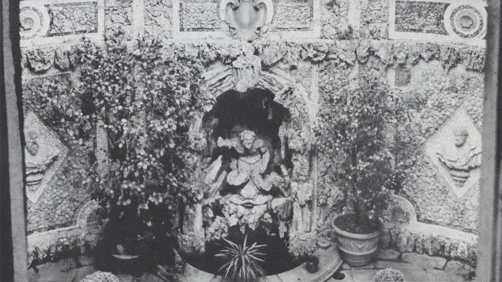 Faded color photo of a yellow wall with a large, grey and white statue of a vase inset near the wall. The vase is surrounded by by green trees with white flowers in an outdoor garden.