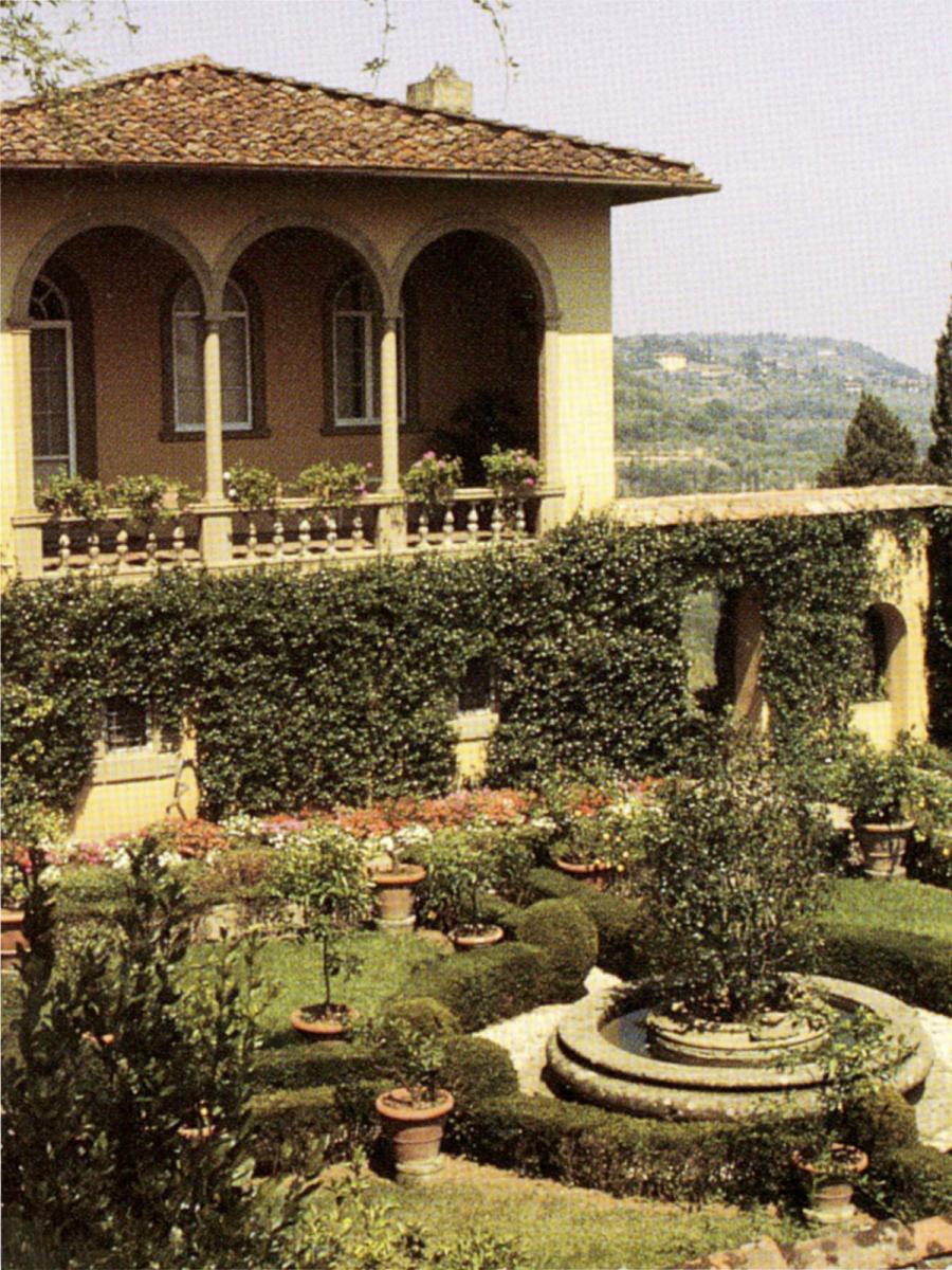 Faded color photo of a walled yellow courtyard, which is divided into four sections by green shrubbery. At the center is a fountain with a tree inside of it. Green vines and pink flowers grow up the sides of the courtyard walls. On the opposite wall, a building with an enclosed balcony overlooks the courtyard. On the right side of the picture, we see that the courtyard sits at high altitude, overlooking the Tuscan countryside and a distant town.