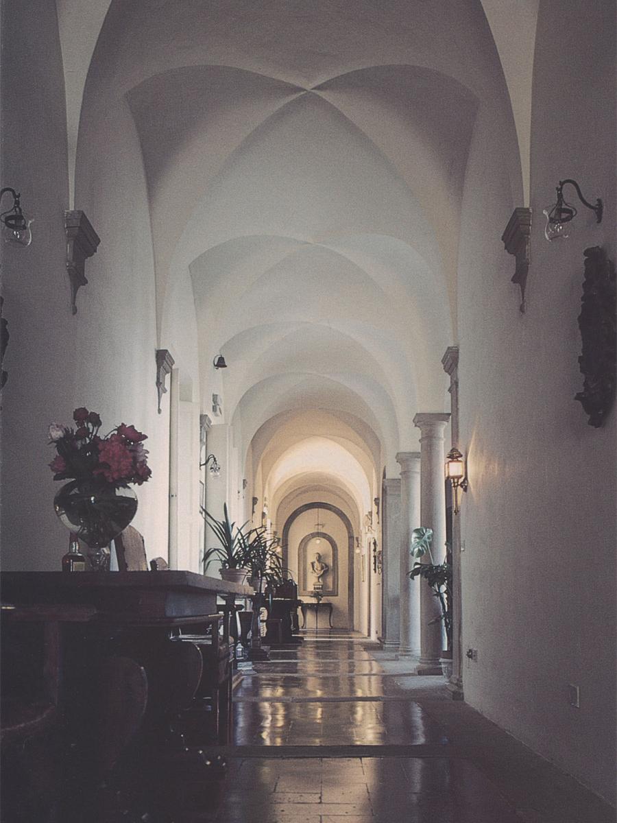 Color photo with an interior view of a long, white hallway with arched ceilings. The hallway is lined with flowers, plants, glass lamps, and old wooden furniture. At the end of the hallway sits a stone bust of a man.